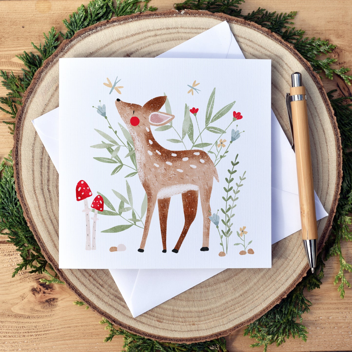 Plain Woodland Deer Birthday Card. Beautifully illustrated deer or fawn, surrounded by green foliage, flowers and toadstools. The card has a white background. Our cards are approximately 6x6 inch and square in shape. We use high quality, textured card for printing to compliment the artwork style. Our cards come with a white envelope and are packaged with biodegradable, eco friendly cellophane for protection from water damage during shipping.